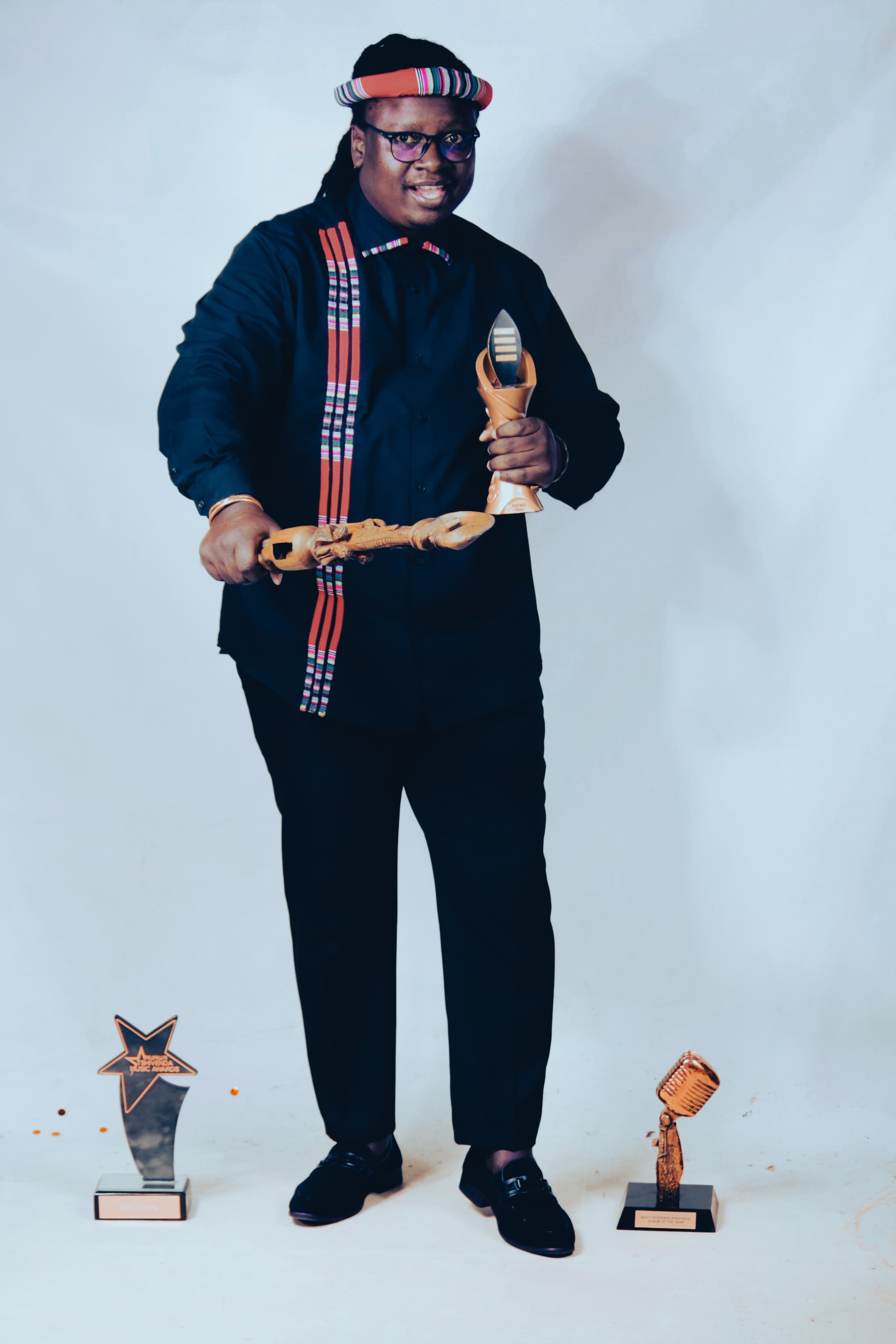 Studio portrait of Papa Thile against a plain pale background, holding a SATMA trophy in one hand and a carved wooden staff in both, wearing a striped Venda headband and sash over black. Two further trophies stand on the floor at either side.