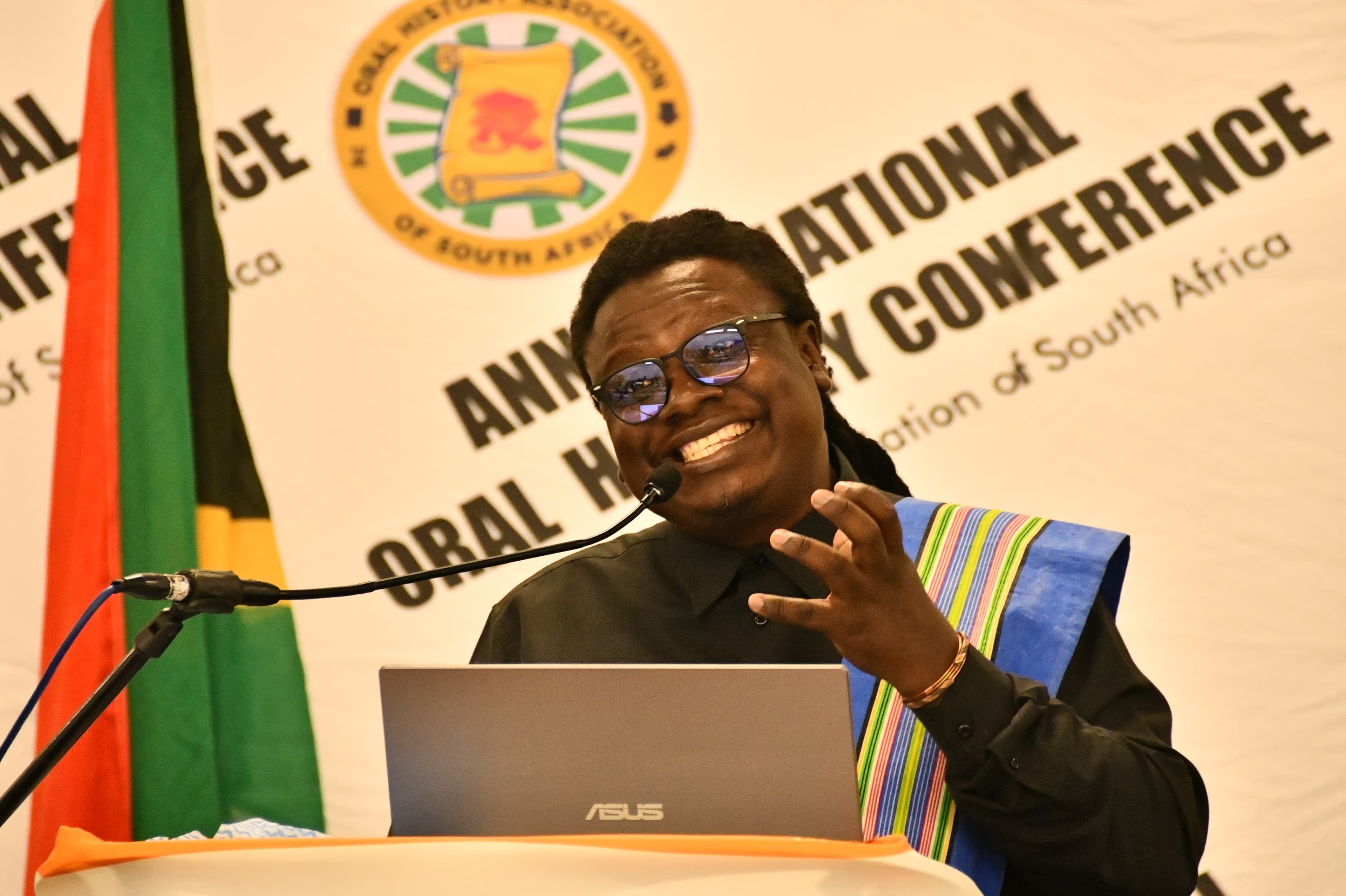 Papa Thile speaking from a lectern at the Annual National Oral History Conference of the Oral History Association of South Africa, mid-sentence and smiling, one hand raised, wearing a woven Venda sash over a dark shirt. A South African flag stands beside the podium.