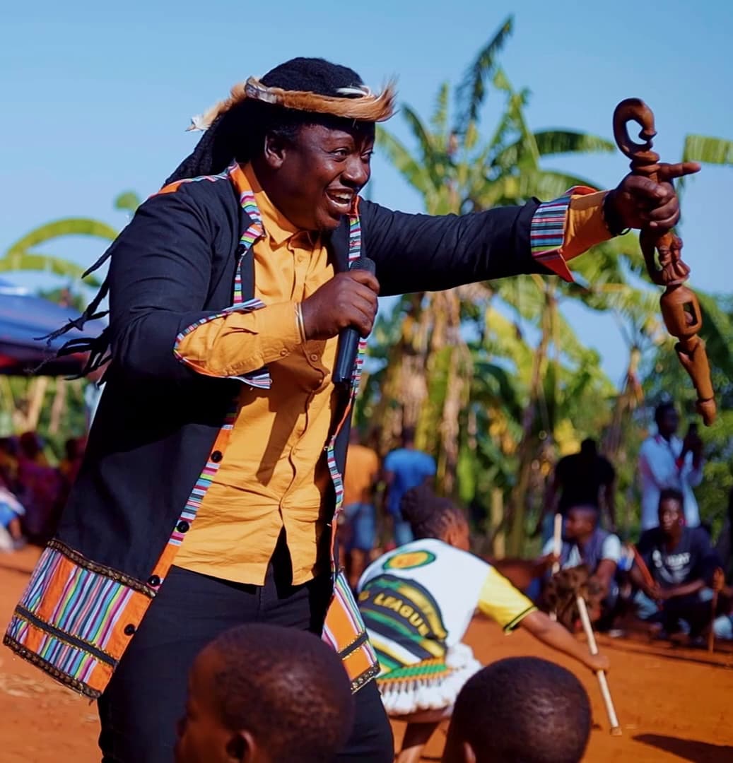 Papa Thile performing outdoors on red earth. Mid-phrase, a microphone in one hand and a carved wooden staff raised in the other, wearing a fur-and-feather headband and a black jacket trimmed with woven Venda cloth. Banana palms, a dancer and a seated audience behind.