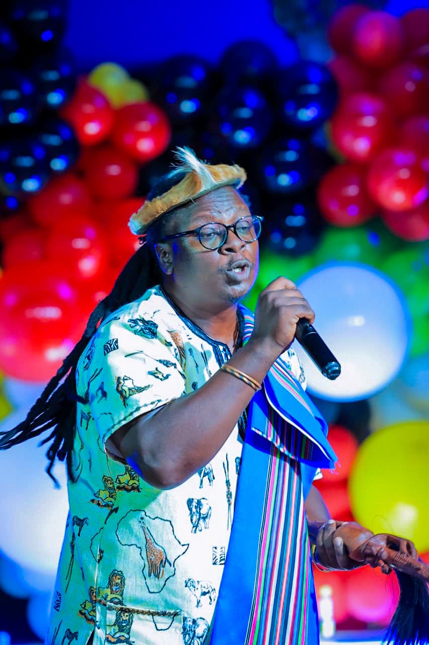 Close stage portrait of Papa Thile singing into a handheld microphone under coloured lights, wearing glasses, a fur headband, and a shirt printed with a map of Africa alongside a woven Venda sash. A wall of dark and red balloons fills the background.