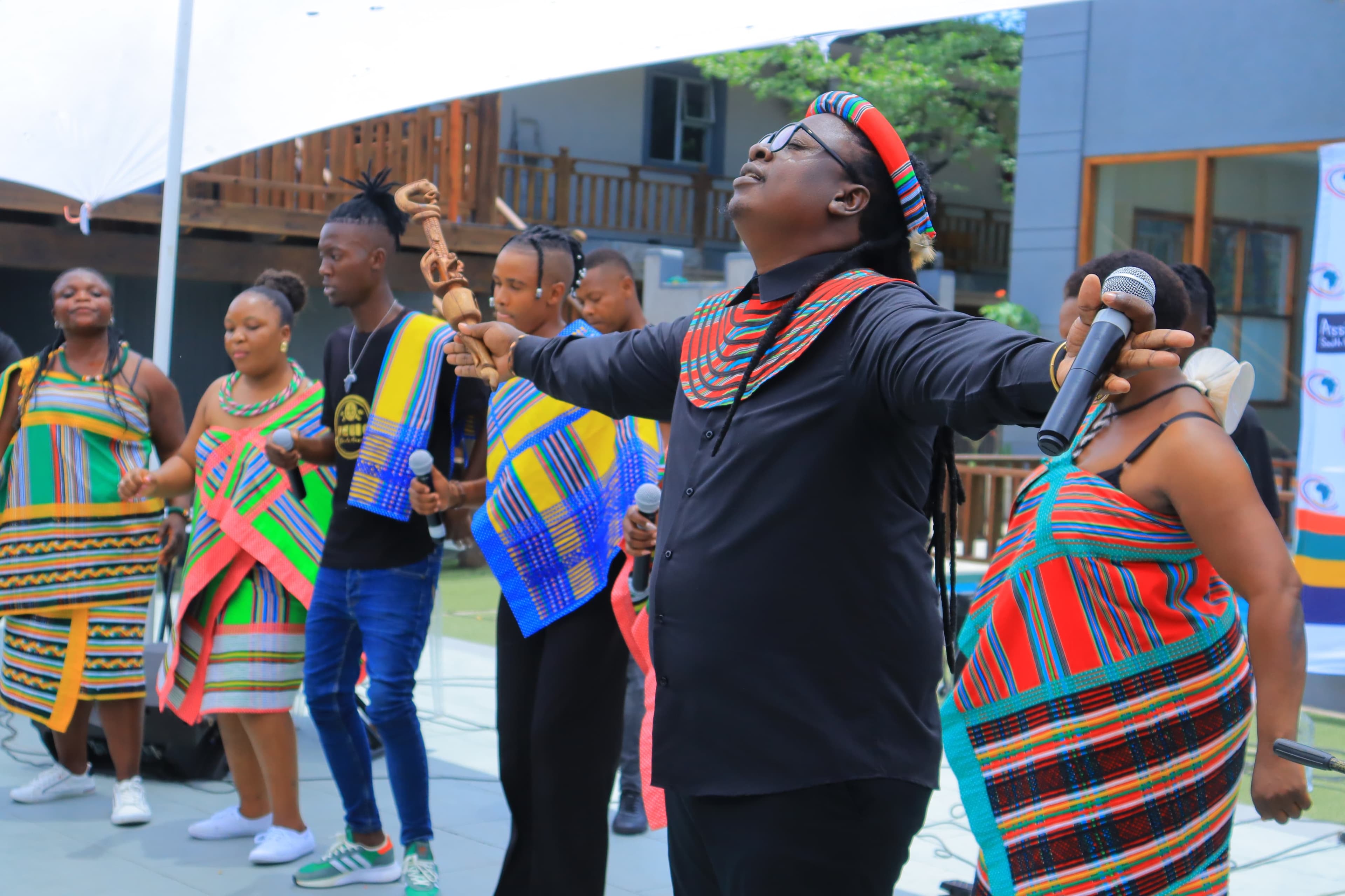Papa Thile performing outdoors with an ensemble. Head tilted back, arms outstretched, a microphone in one hand and a carved wooden staff in the other, wearing a striped Venda headband and collar. Performers in Venda dress stand around, several holding microphones.